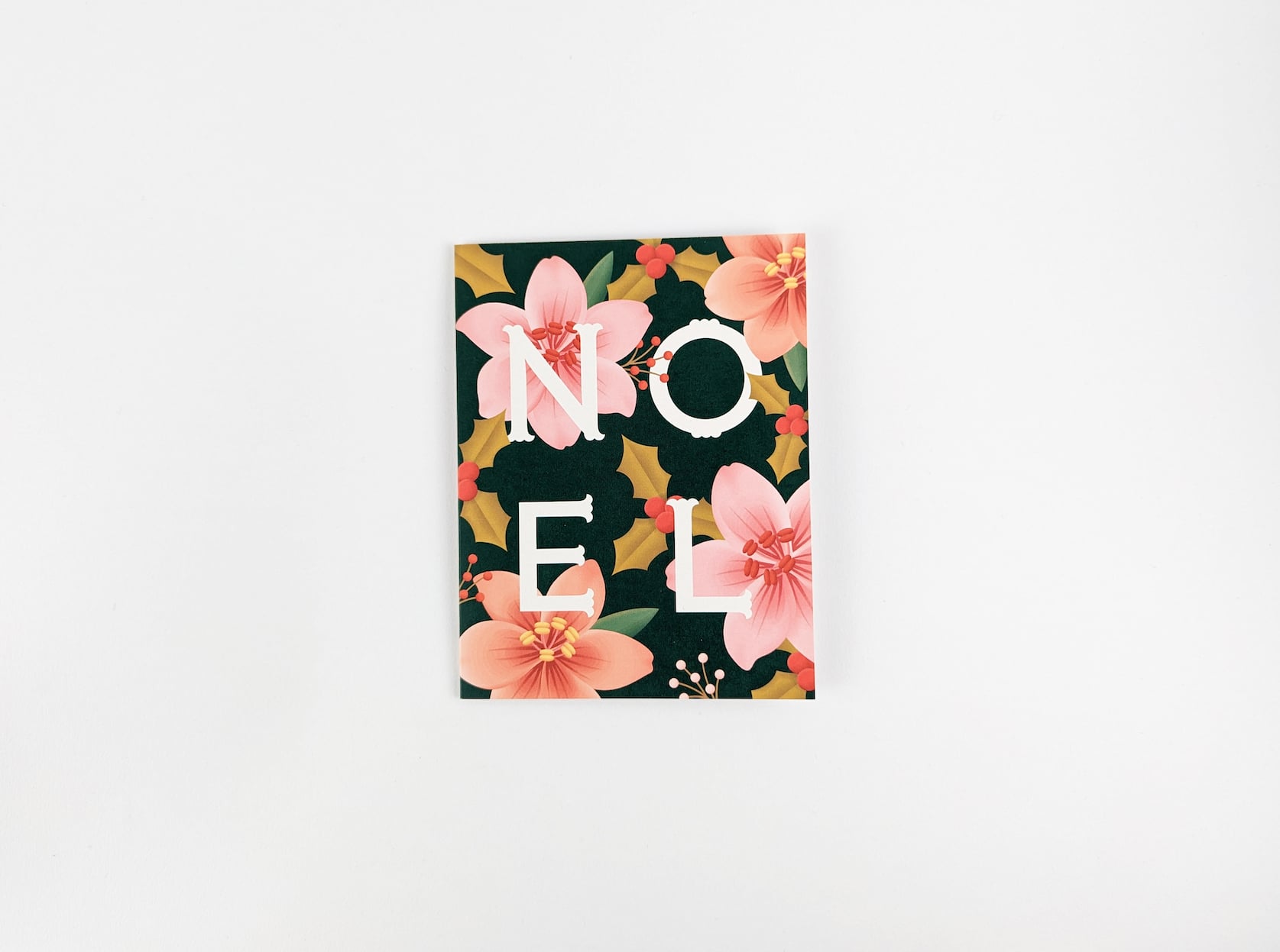 Dark green card with white text in the centre that reads: Noel. Pink amaryllis flowers are featured around the text.