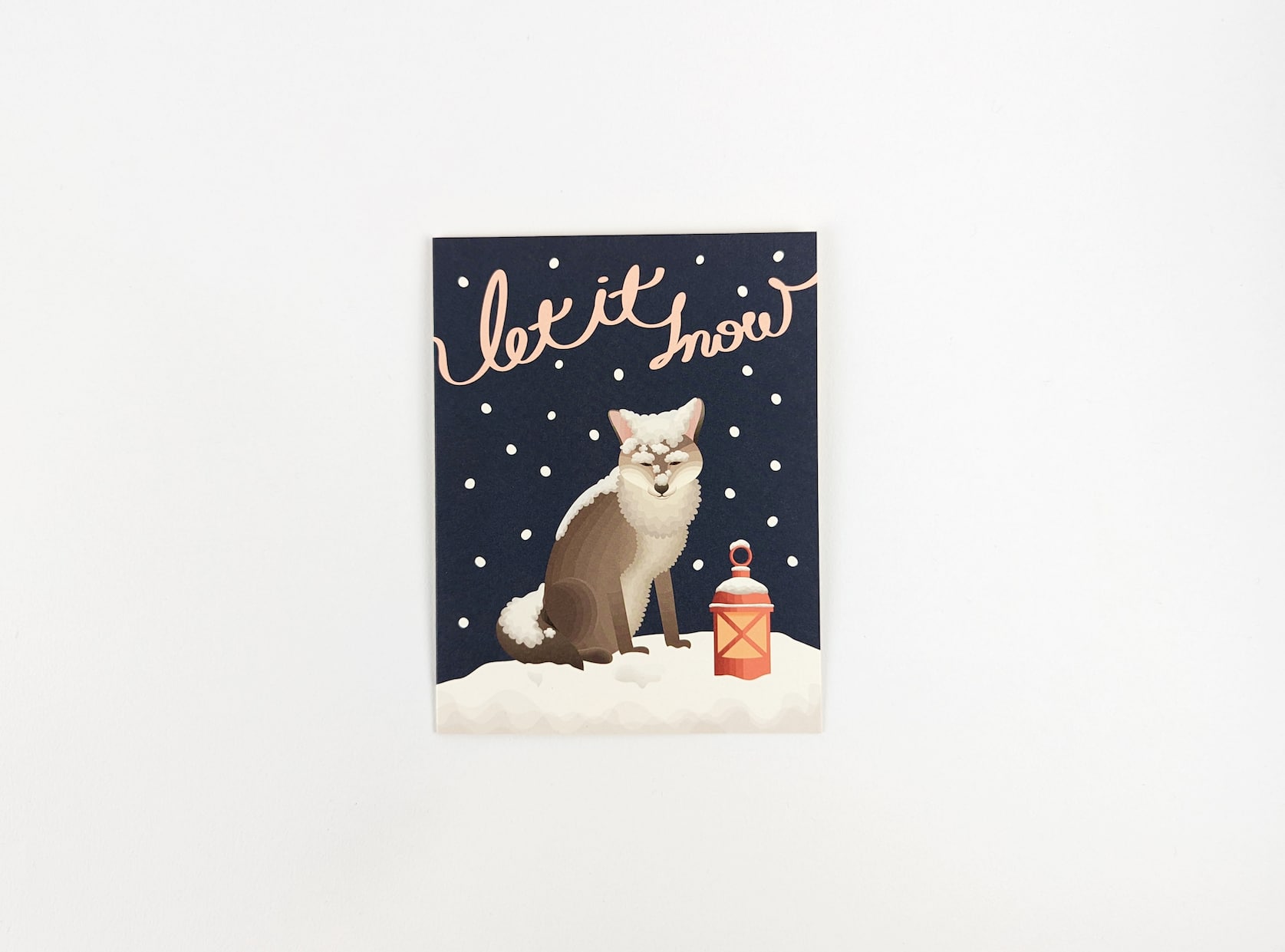 Pink cursive text reads: Let it snow. Below a brown fox stands in the snow with a red lantern beside him. The background is dark blue with snow falling around.