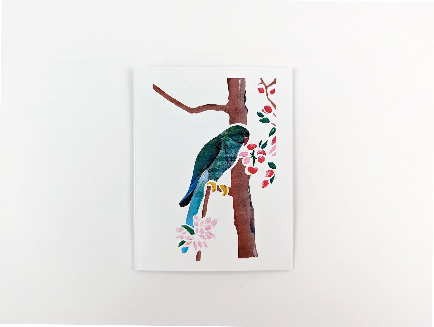 White card with a painted green & blue parrot perched on a branch. Pink & red berries and flowers hang beside the parrot and a pink flower below