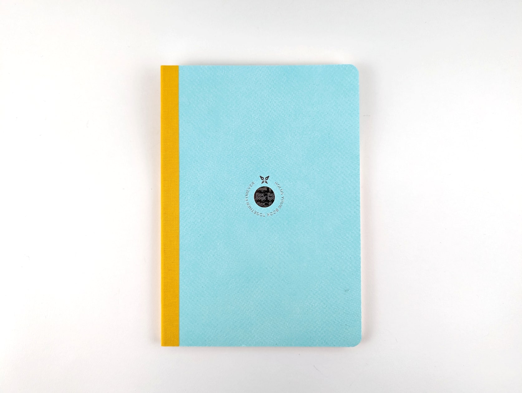 A notebook with light blue-green eco paper and a yellow linen spine. A circular holographic logo is featured in the center with a butterfly and silver text wrapping around that reads: Holds your book together forever.