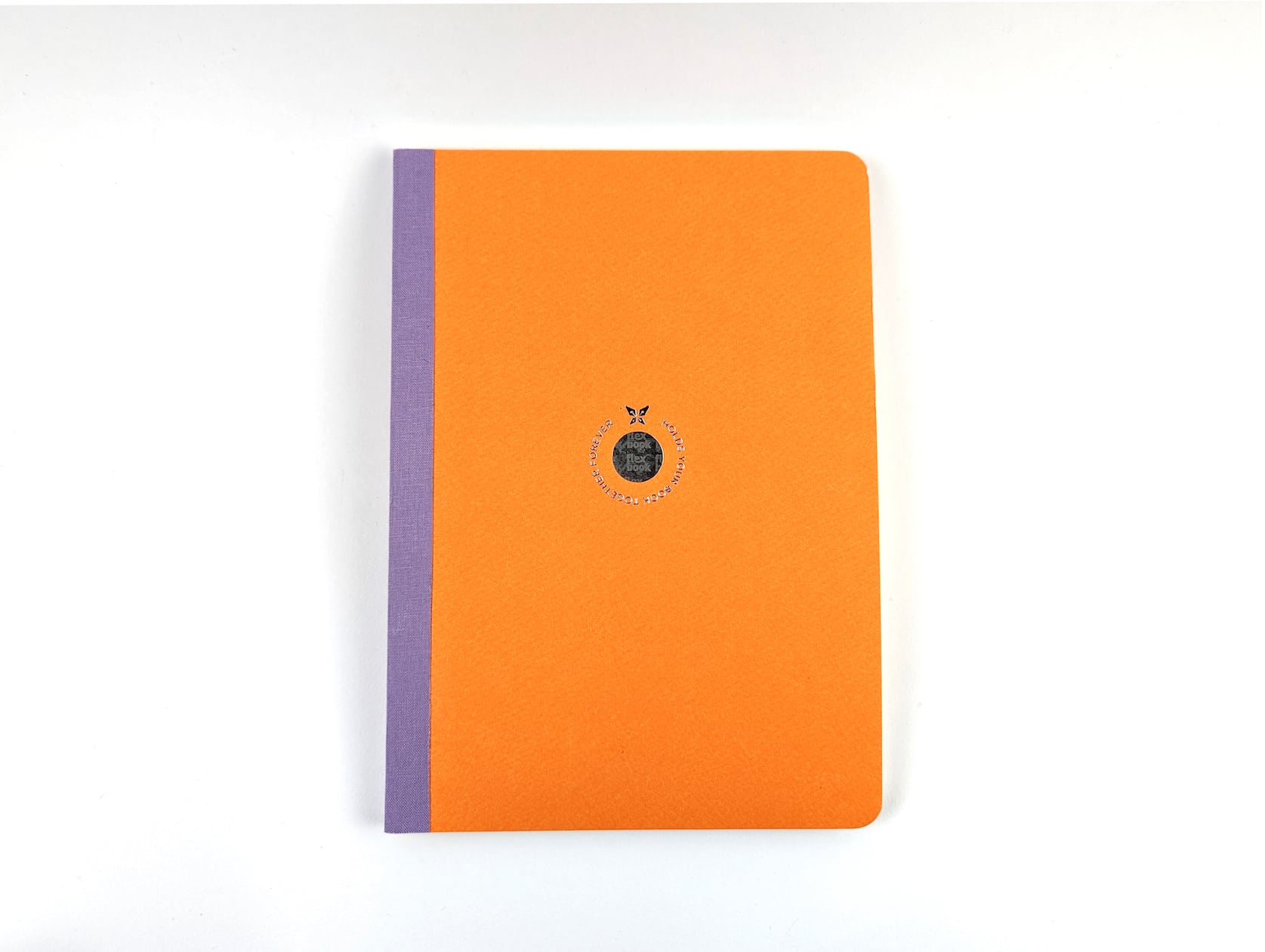 A notebook with orange eco paper and a purple linen spine. A circular holographic logo is featured in the center with a butterfly and silver text wrapping around that reads: Holds your book together forever.