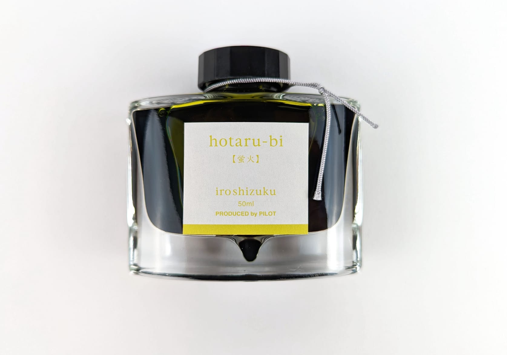 A rectangular glass ink bottle with a silver string tied around its neck. Square sticker with yellow-green text that reads: hotaru-bi. iroshizuku. 50 ml. Produced by Pilot.