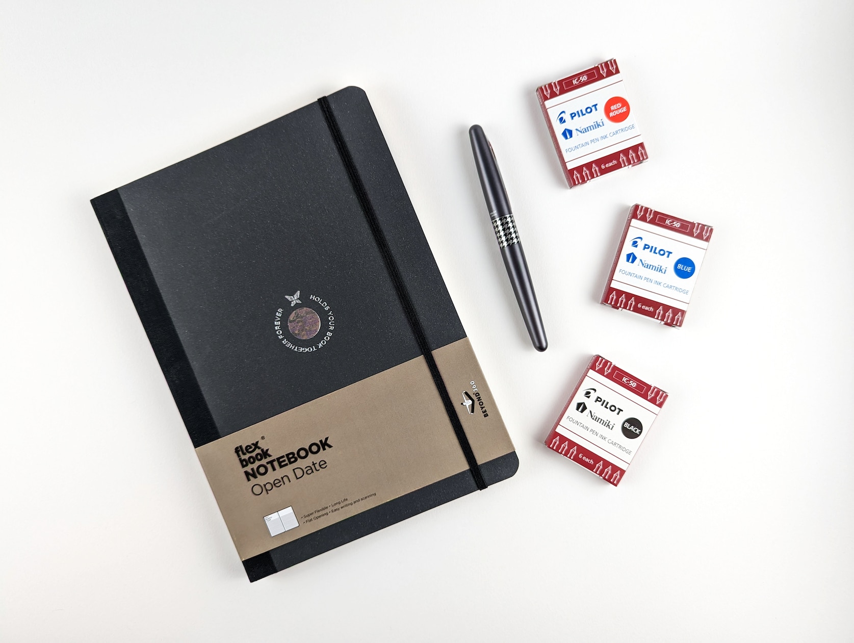 Bundle of items on a white background. Items include: black notebook, houndstooth black fountain pen and three packs of fountain pen ink in three different colours: black, blue and red.