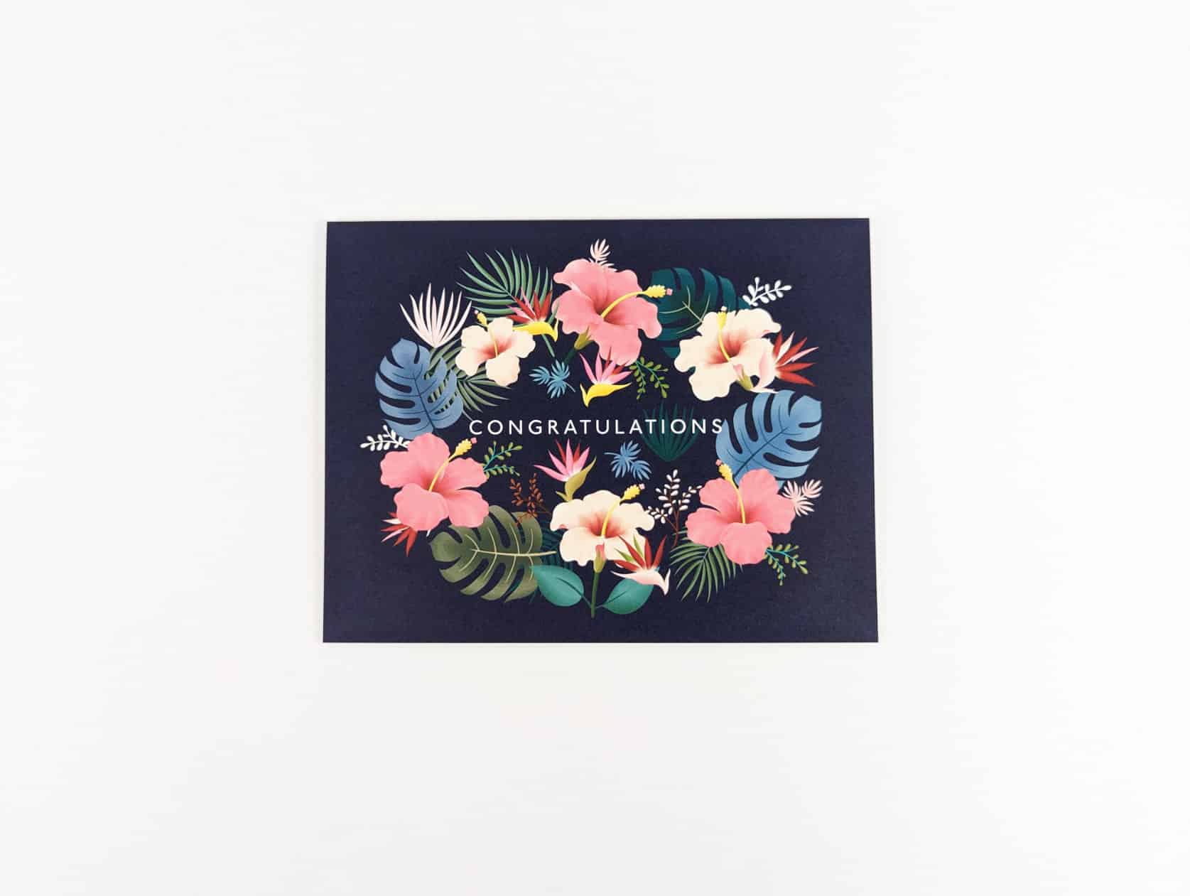 Navy horizontal card with white text in the centre that reads: CONGRATULATIONS. Hibiscus flowers, green & blue monstera leaves and white, green and brown leaves surround the text in a ring.