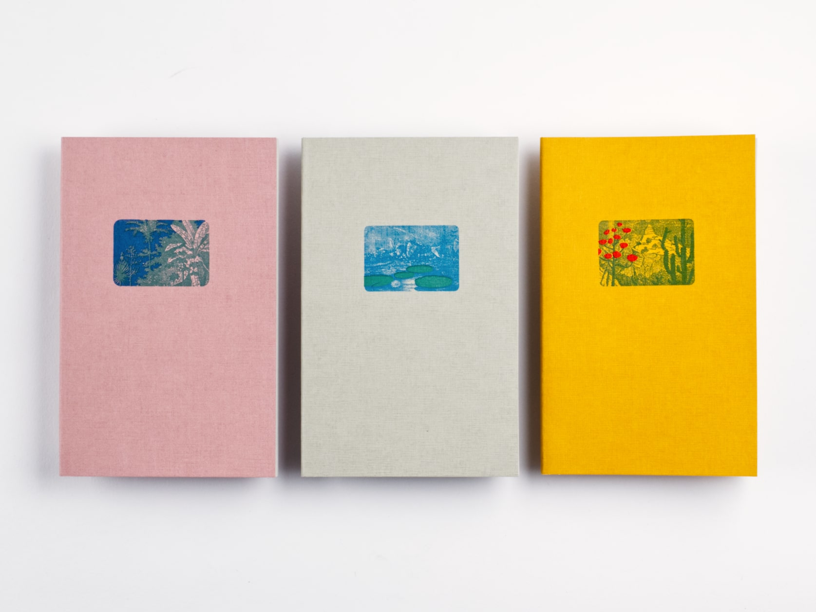 3 softcover notebooks positioned side by side. One pink with a print of palm trees, one green with a print of lilypads and one yellow with a print of a cactus beside flowers