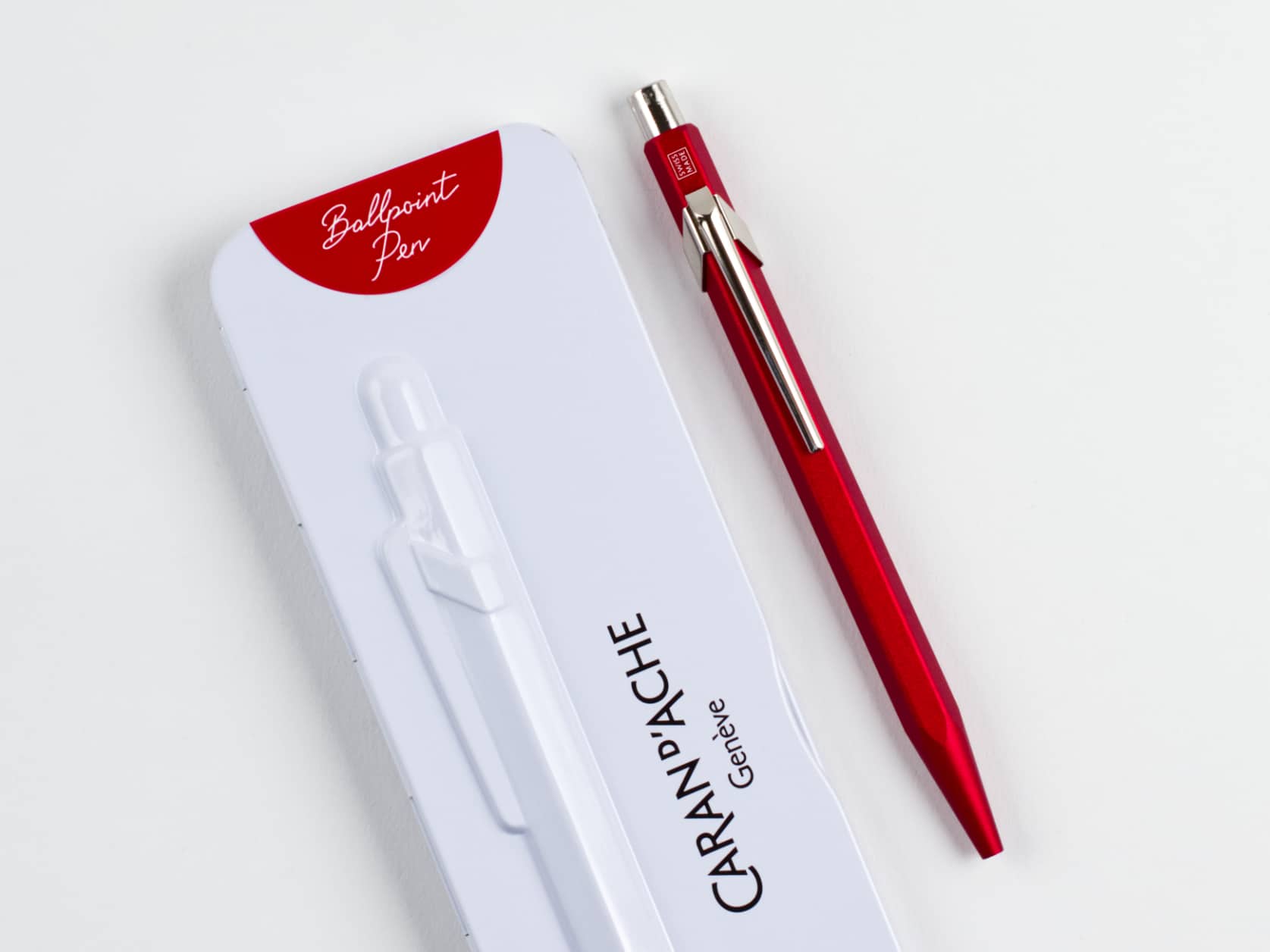 Metal ballpoint pen with a matte red hexagonal barrel and silver-coloured metal clip and cap. Beside it, its white metal case which has a red semi-circle at one end containing the text: Ballpoint Pen. The case also has a relief of the pen and the Caran d'Ache logo.