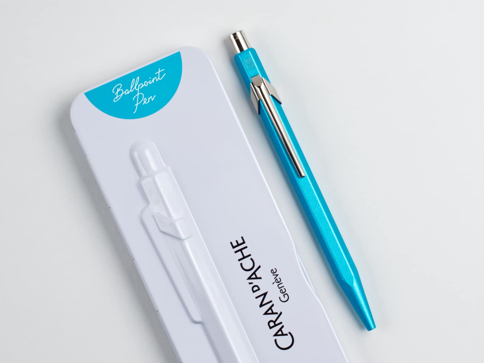 Metal ballpoint pen with a blue matte hexagonal barrel and silver-coloured metal clip and cap. Beside it, its white metal case which has a blue semi-circle at one end containing the text: Ballpoint Pen. The case also has a relief of the pen and the Caran d'Ache logo.