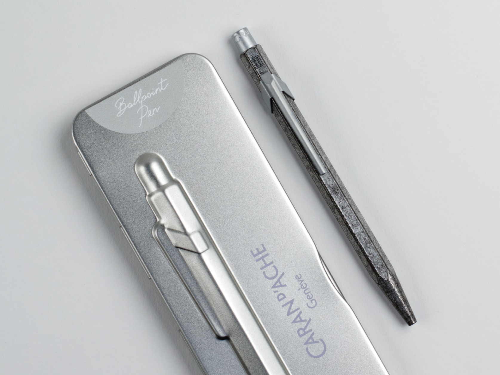 Metal ballpoint pen with aged metal coloured hexagonal barrel and silver-coloured metal clip and cap. Beside it, its grey metal case which has a grey semi-circle at one end containing the text: Ballpoint Pen. The case also has a relief of the pen and the Caran d'Ache logo.