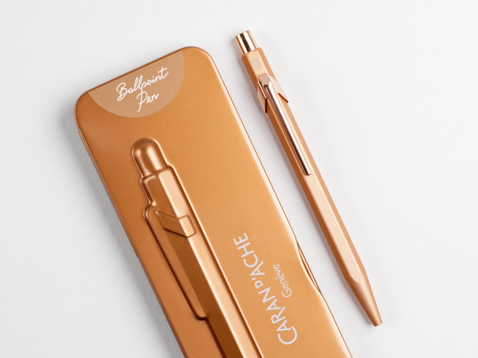 Metal ballpoint pen with a matte orange hexagonal barrel and copper-coloured metal clip and cap. Beside it, its orange metal case which has semi-circle at one end containing the text: Ballpoint Pen. The case also has a relief of the pen and the Caran d'Ache logo.