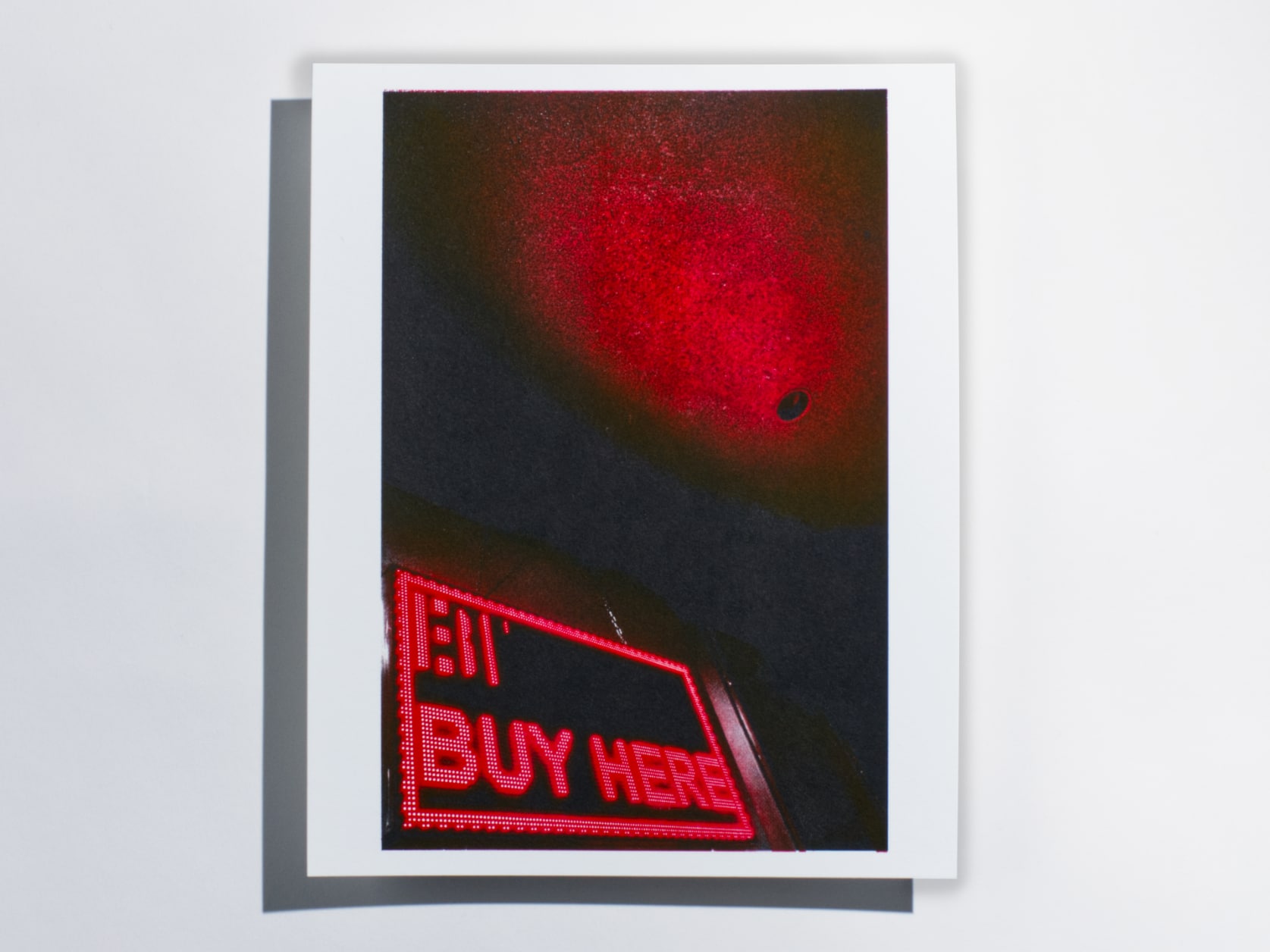 Photography print of a dark room. The ceiling is reflecting the light from a large red LED sign spelling out the words BUY HERE.