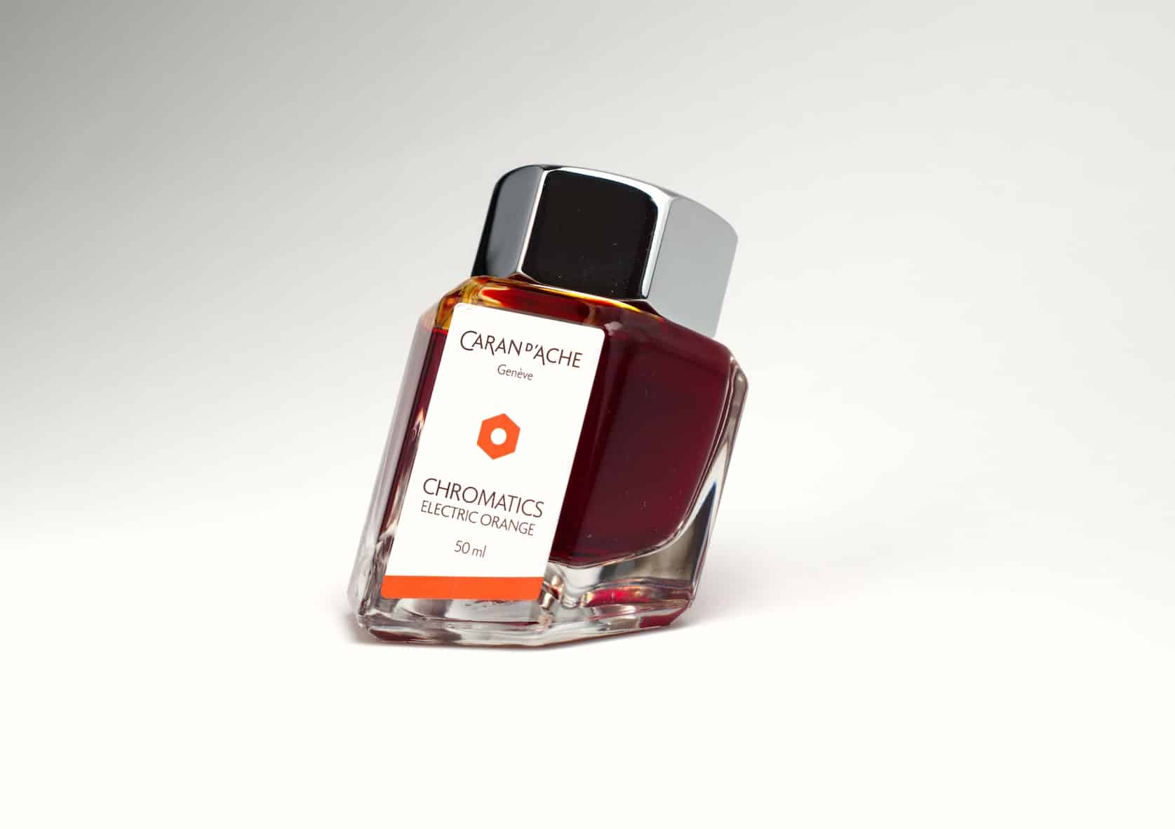 Hexagonal ink bottle with a metal cap and a base skewed on an angle. The bottle contains orange ink and has a label with the Caran d'Ache logo. Text reads: Chromatics, Electric Orange, 50 ml.