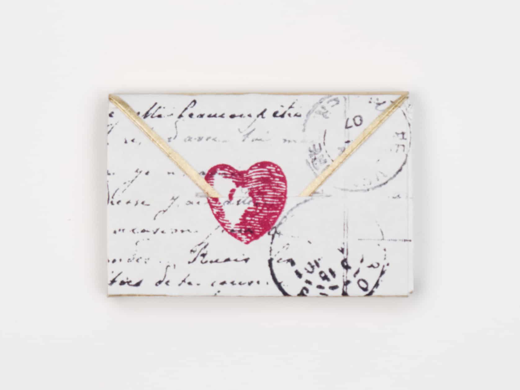 A mini envelope with gold trim on the flap, some writing and stamping on the front and a stamped red heart in the middle.