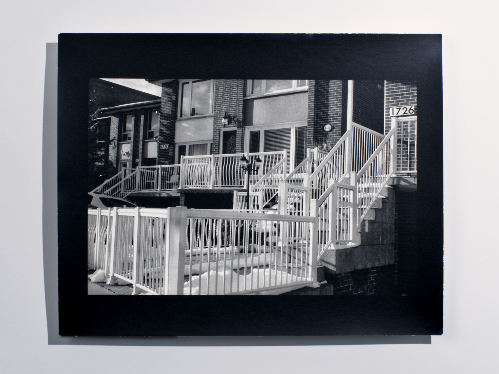 A black and white photography print of the front of a house gated by a long fence