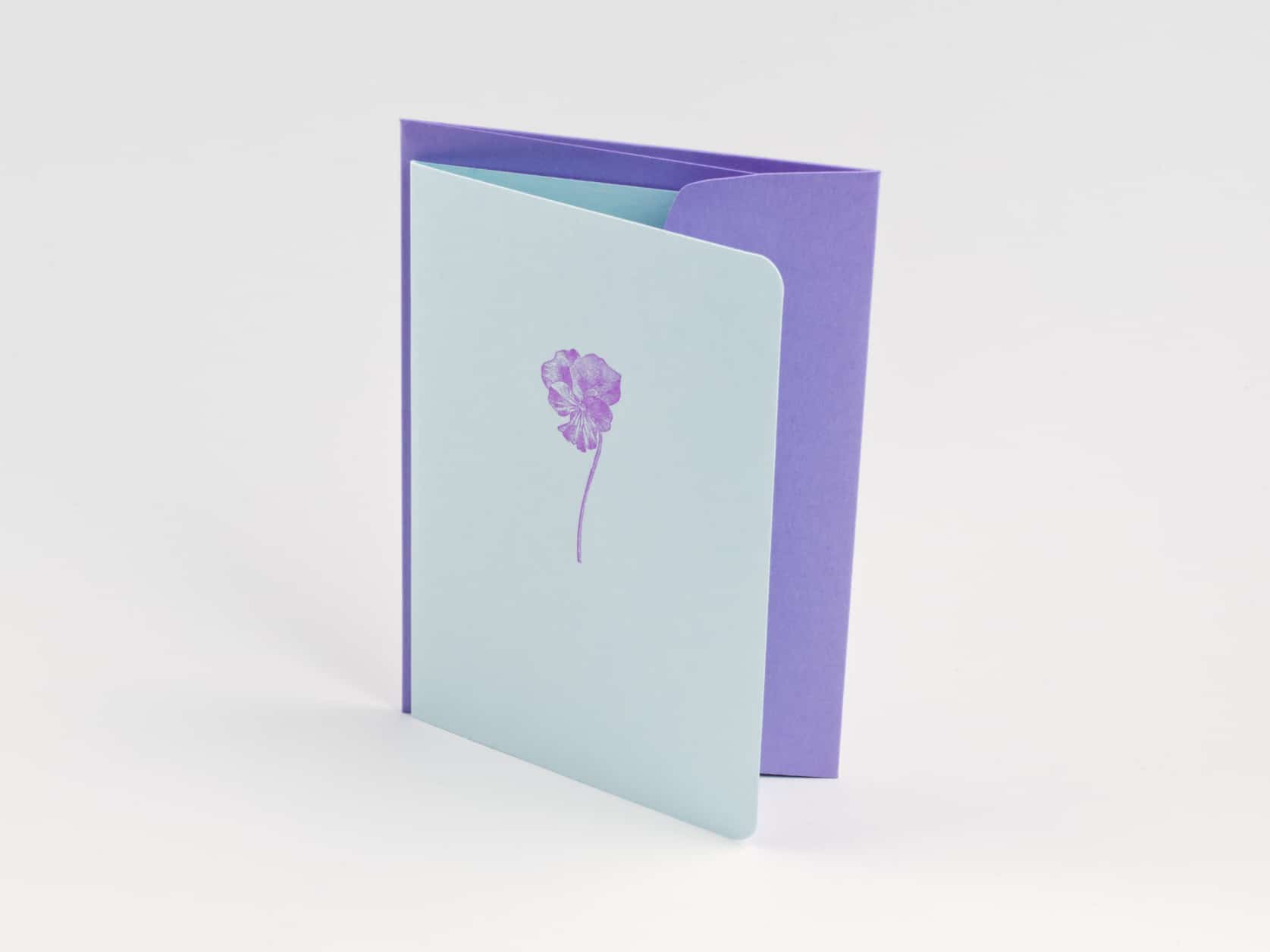 A blue card with an illustration of a purple pansy flower on it and a purple envelope standing upright tightly together