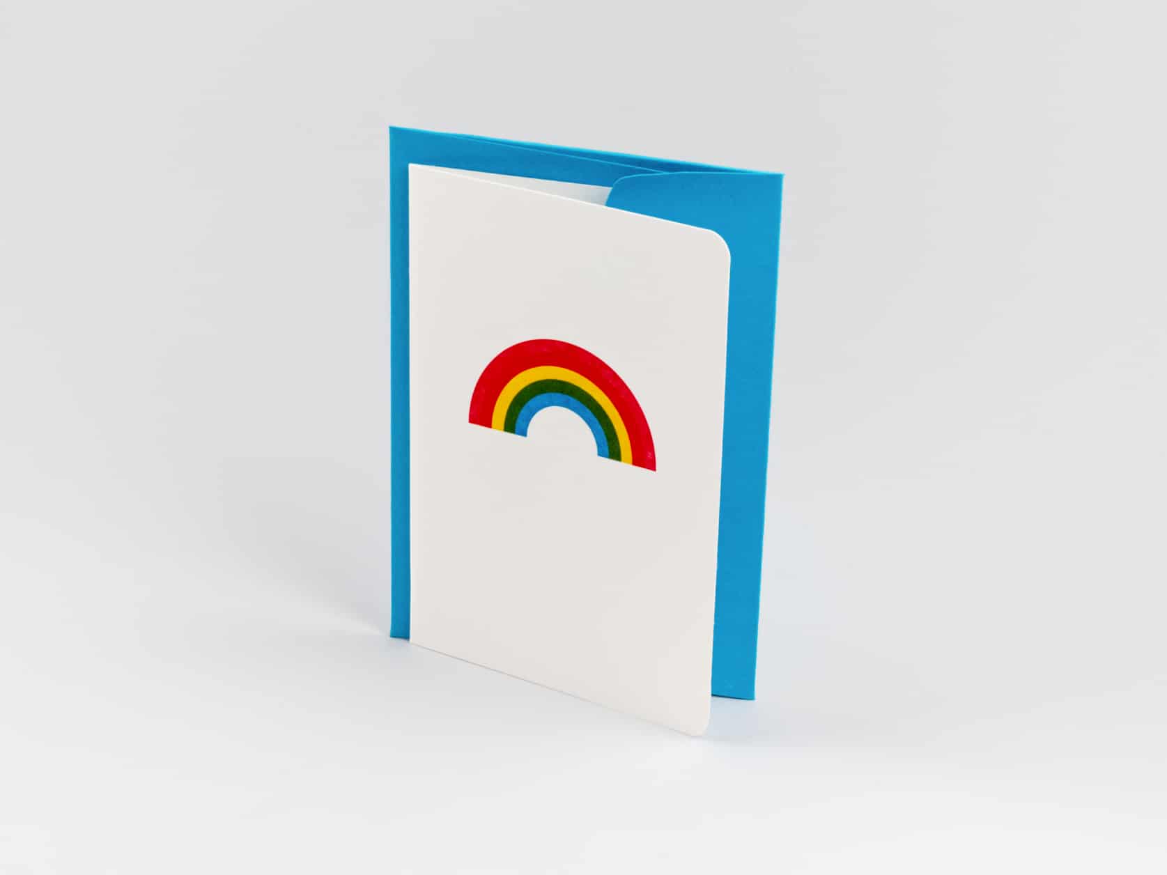 A white card with an illustration of a rainbow on it and a blue envelope standing upright tightly together