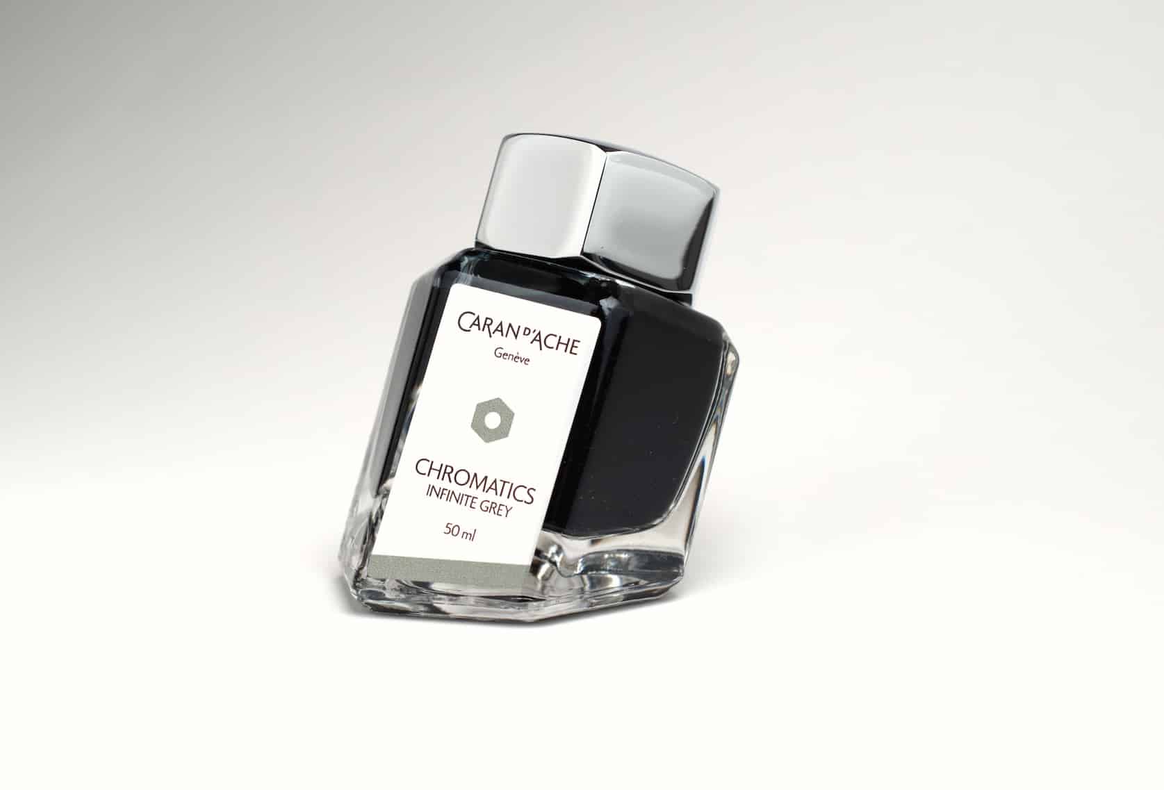 Hexagonal ink bottle with a metal cap and a base skewed on an angle. The bottle contains black ink and has a label with the Caran d'Ache logo. Text reads: Chromatics, Infinite Grey, 50 ml.