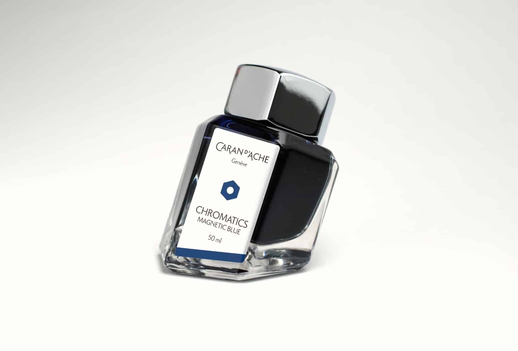 Hexagonal ink bottle with a metal cap and a base skewed on an angle. The bottle contains black ink and has a label with the Caran d'Ache logo. Text reads: Chromatics, Magnetic Blue, 50 ml.