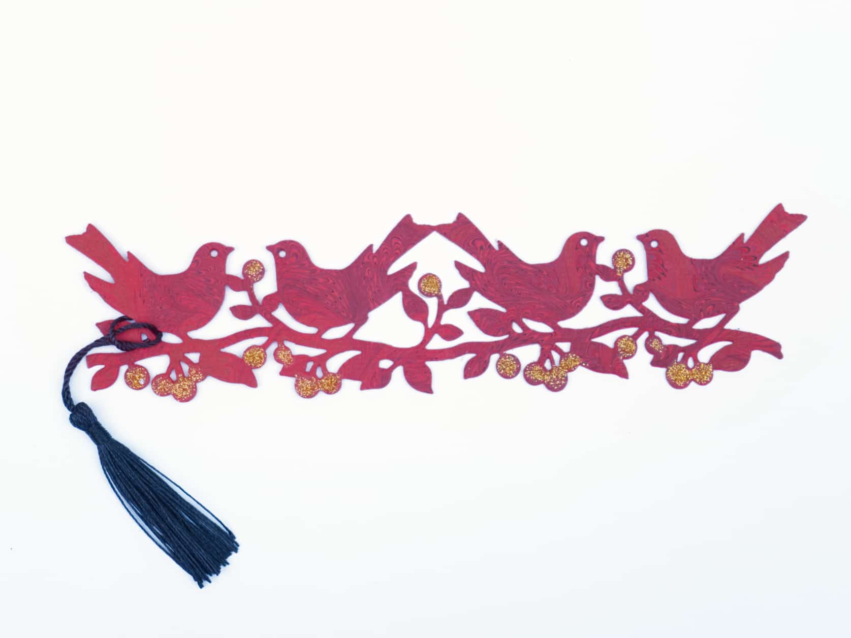 Bookmark consisting of a cutout of four birds on a branch with bulbs coated in gold glitter and a black tassel on the end. The paper has an intricate red, black and purple marbled design.