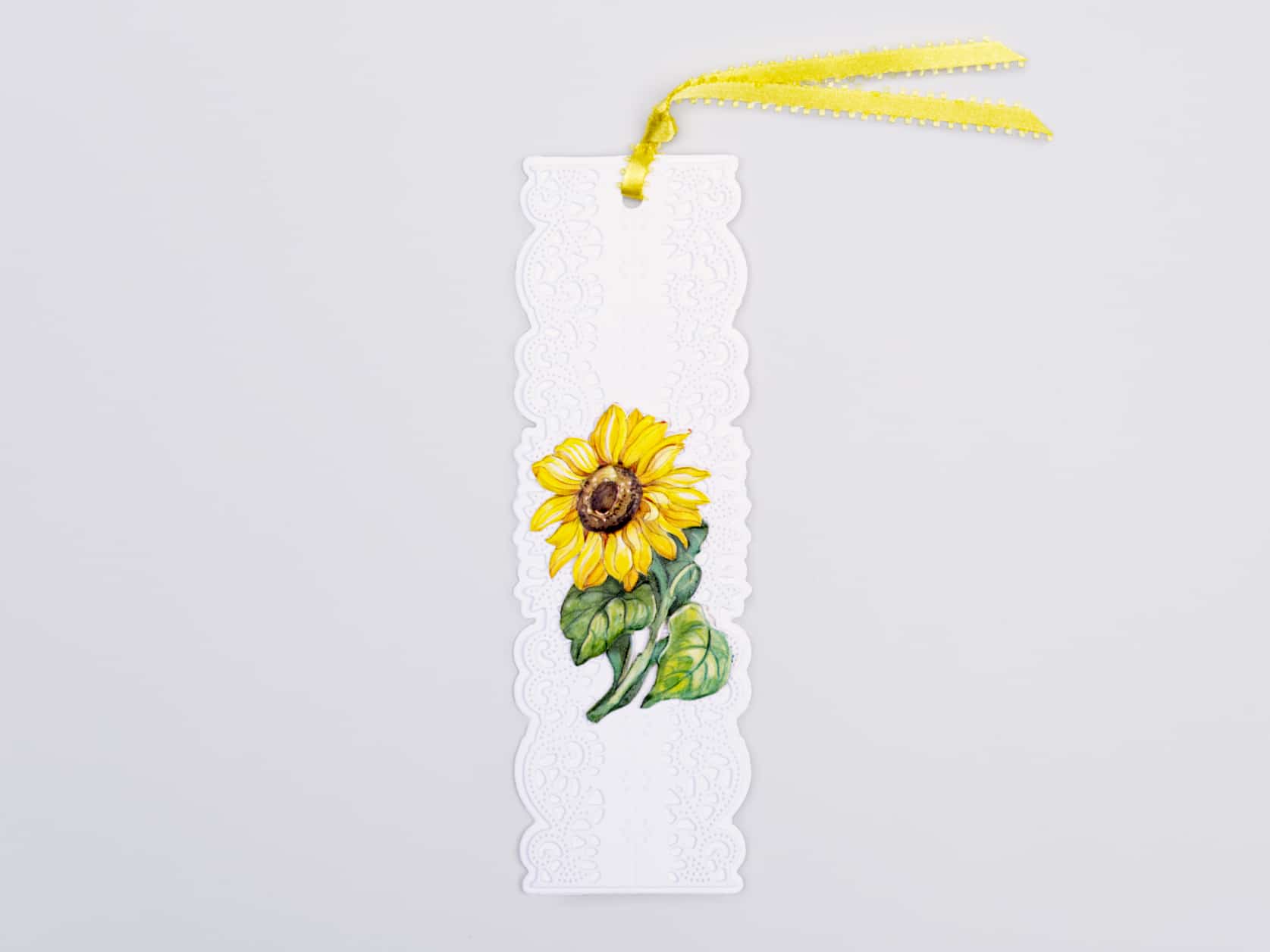 White paper lace bookmark with an illustration of a sunflower in the middle and a yellow ribbon at the top