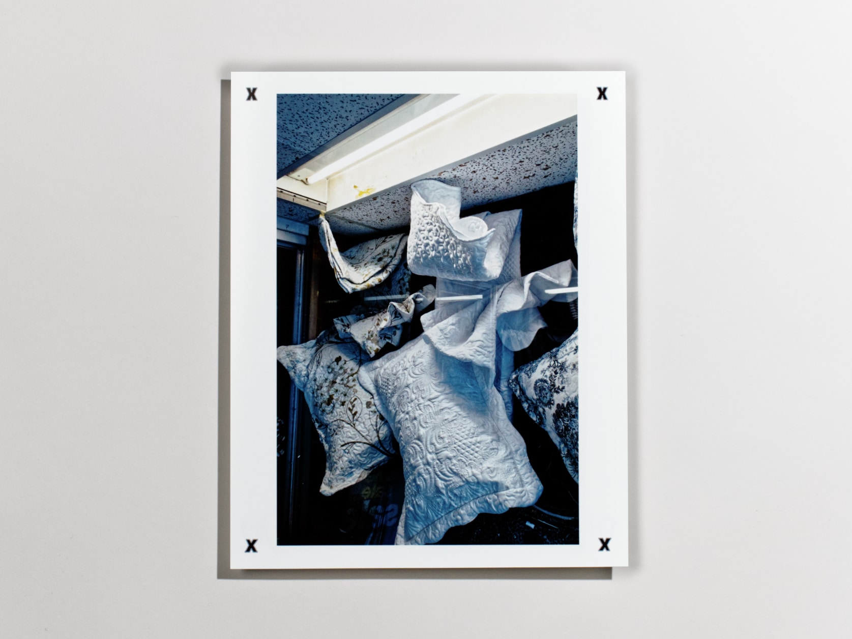 Photography print of florally textured white pillows hanging under a fluorescent light bar in a dark shop window.