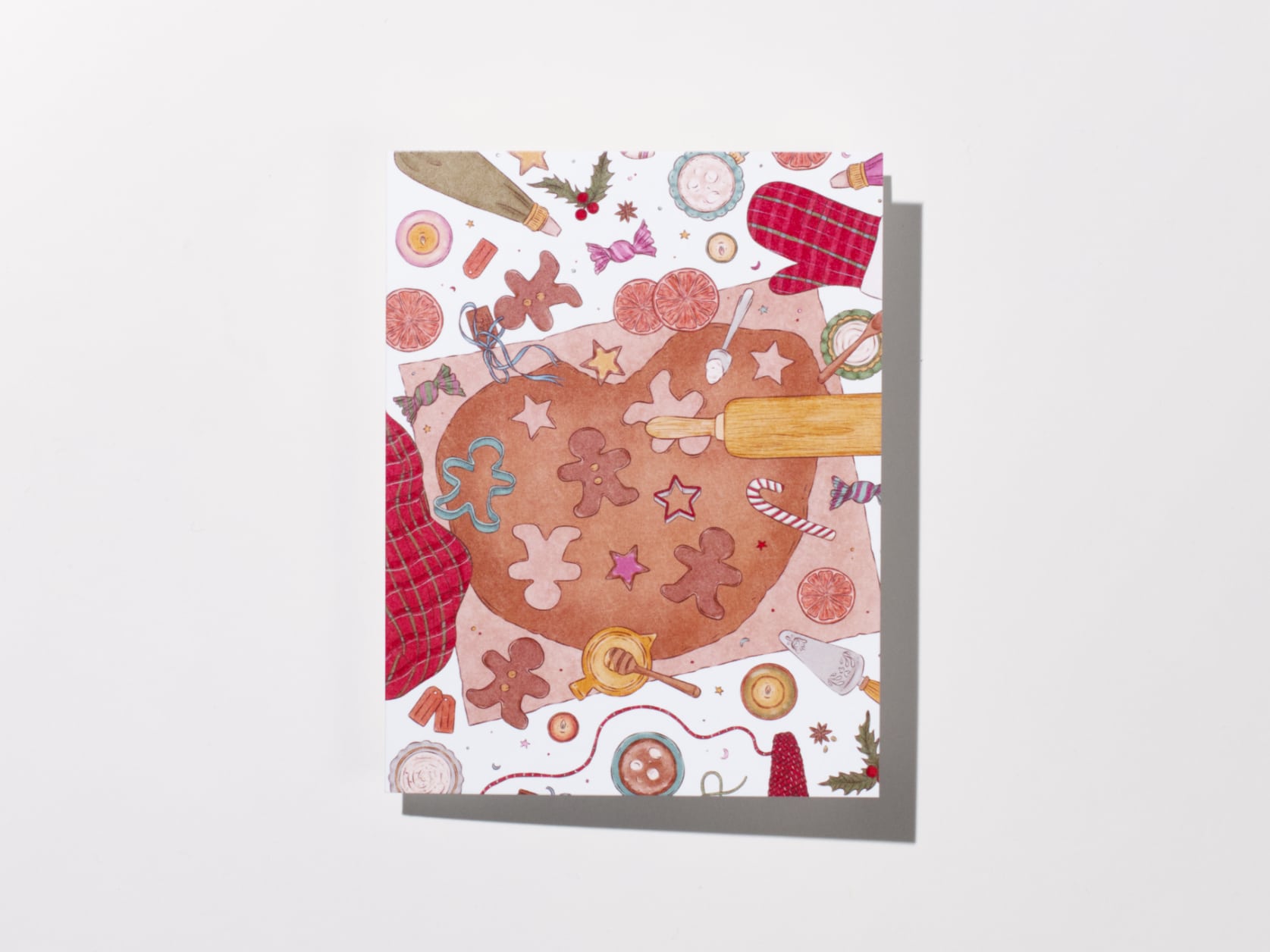 A greeting card featuring cookie dough rolled out on a countertop with various cookie shapes, oven gloves and a rolling pin nearby.